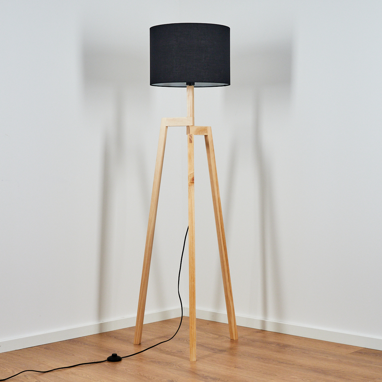 Eine hölzerne Stehlampe mit schwarzem Lampenschirm steht vor einer weißen Wand auf Holzboden.