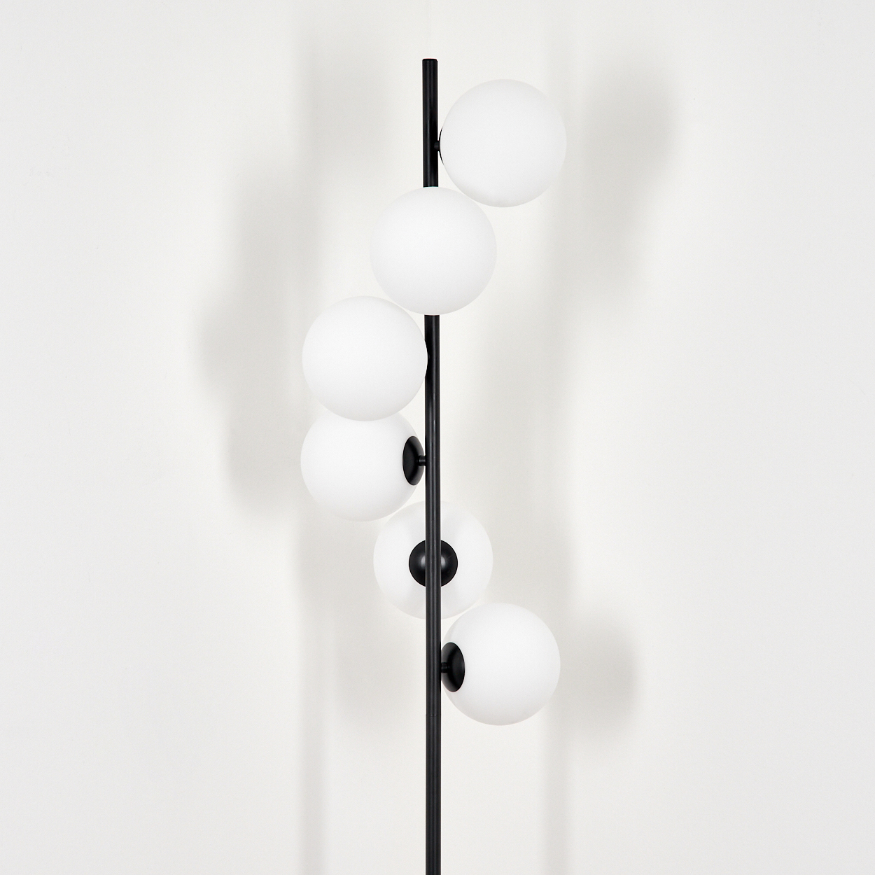 Eine schwarze Stehlampe mit sechs weißen Kugelleuchten steht vor einer weißen Wand. Die Lampe wirft einen Schatten auf die Wand.