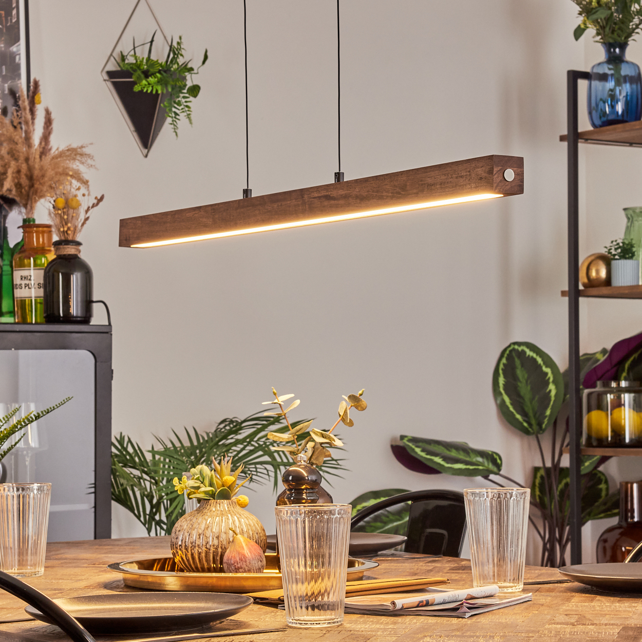 Ein Esszimmer mit einem Holztisch, der für eine Mahlzeit gedeckt ist. Eine Holzlampe hängt darüber. Dekorative Pflanzen und Regale sind im Hintergrund.
