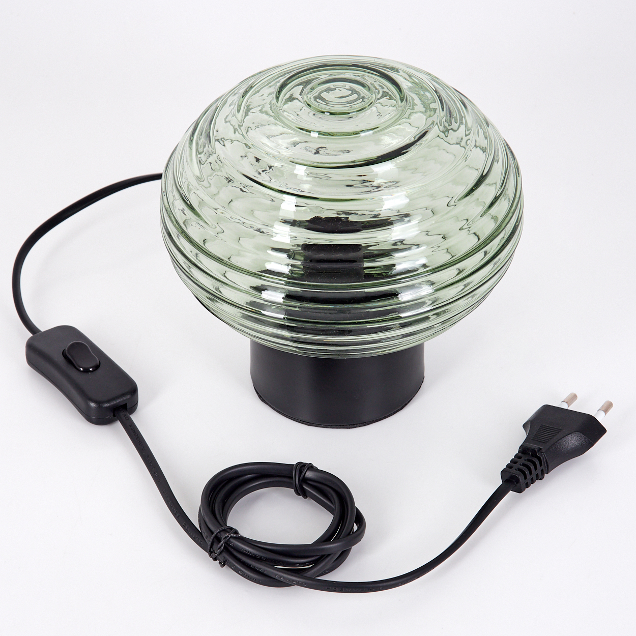 Eine Glaslampe mit grün getöntem, geripptem Globus, der auf einem schwarzen Sockel ruht. Ein schwarzes Kabel mit Schalter und Stecker erstreckt sich von der Lampe.