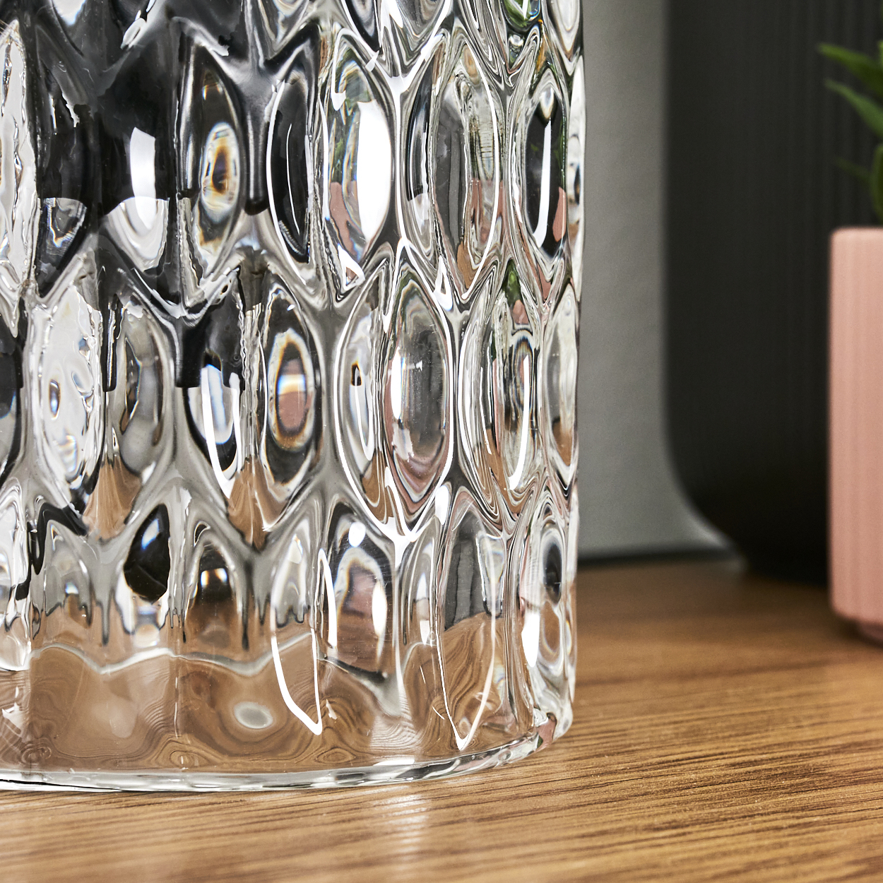 Nahaufnahme eines transparenten Glasobjekts mit strukturierter Oberfläche. Das Glas steht auf einer Holzoberfläche. Dunkler Hintergrund, rosa Topf.