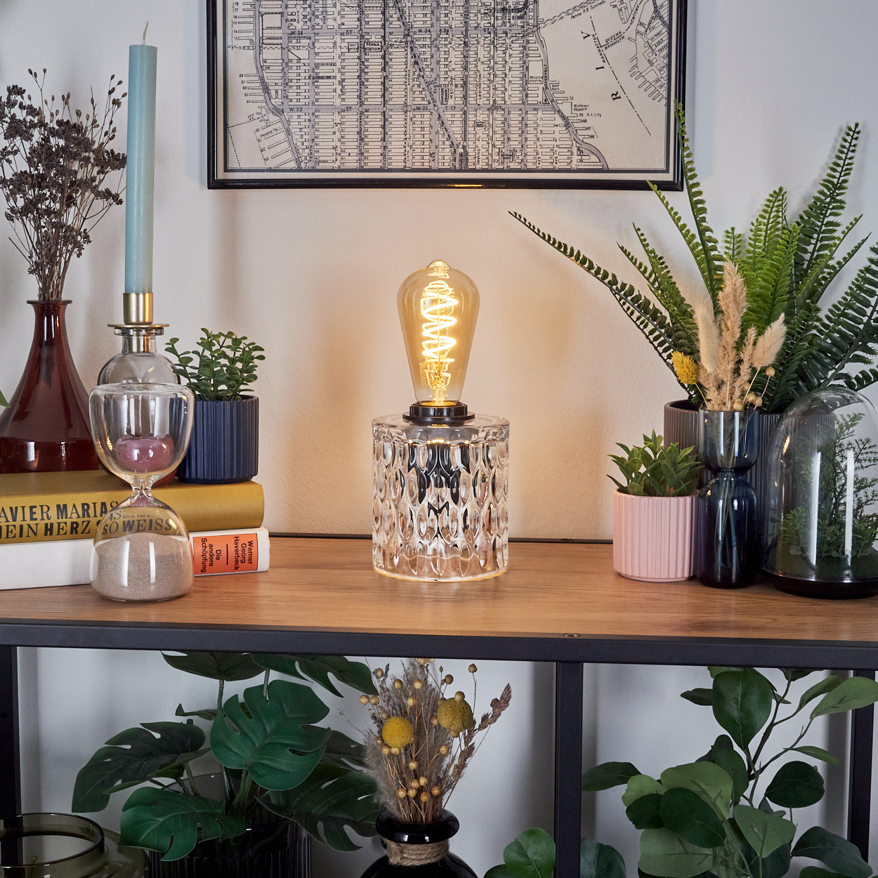 Ein dekoratives Regal mit Lampe, Pflanzen und Karte. Eine gläserne Sanduhr, eine Kerze und Bücher sind ebenfalls vorhanden.