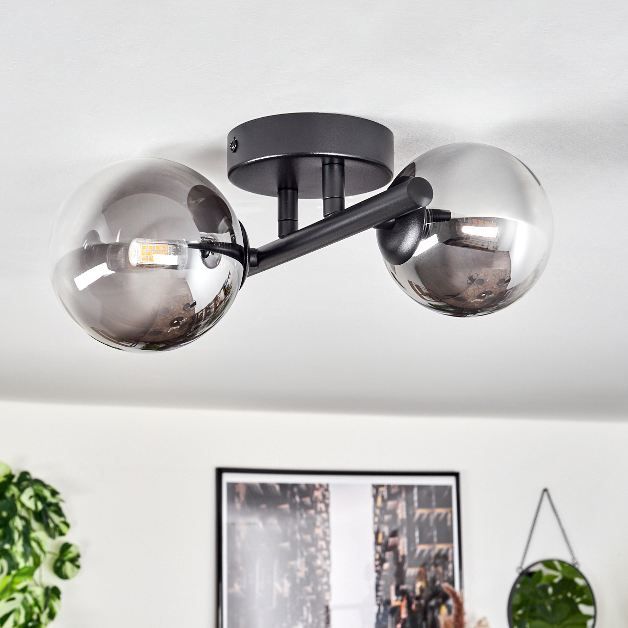 Eine schwarze Deckenlampe mit zwei Glaskugeln. Der Hintergrund zeigt eine weiße Decke und einen Bilderrahmen.