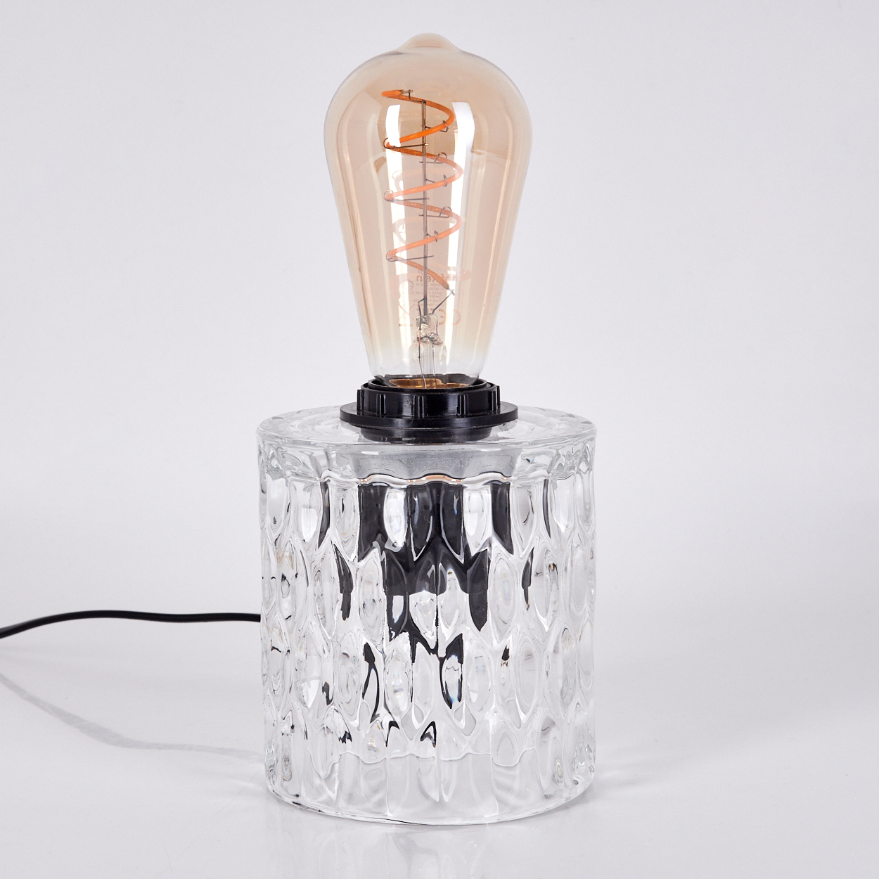 Eine dekorative Tischlampe. Sie hat einen Sockel aus klarem, strukturiertem Glas. Die Glühbirne ist im Vintage-Stil mit orangefarbenem Wendel.