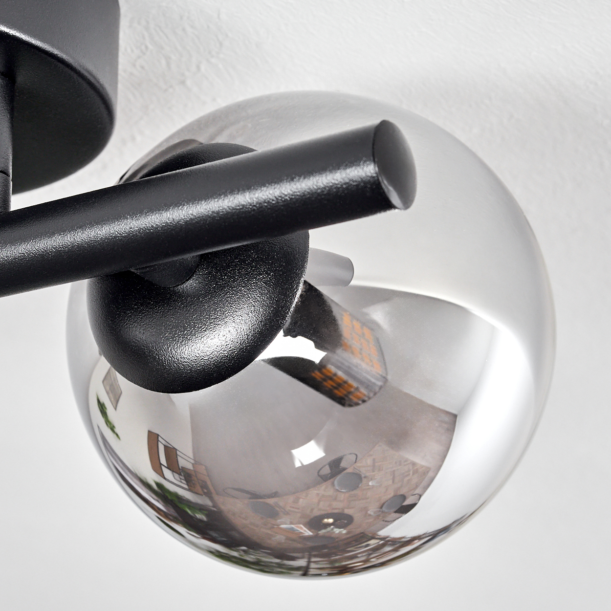 Eine moderne Deckenlampe mit einer reflektierenden Glaskugel. Die Kugel spiegelt einen Raum mit Tischen und Stühlen wider. Schwarze Metallarme und -basis.