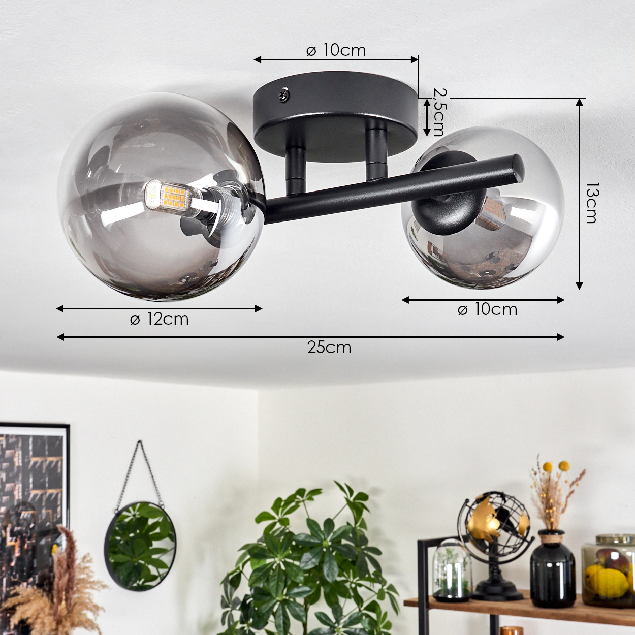 Eine schwarze und graue Deckenlampe mit zwei Glaskugeln, Maße angegeben. Ein Spiegel und Pflanzen sind im Hintergrund.