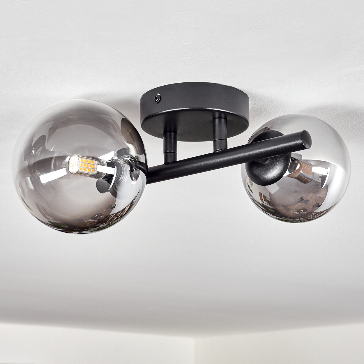Deckenleuchte mit zwei rauchfarbenen Glaskugeln und schwarzem Metallrahmen, an einer weißen Decke montiert. Eine Glühbirne ist sichtbar.