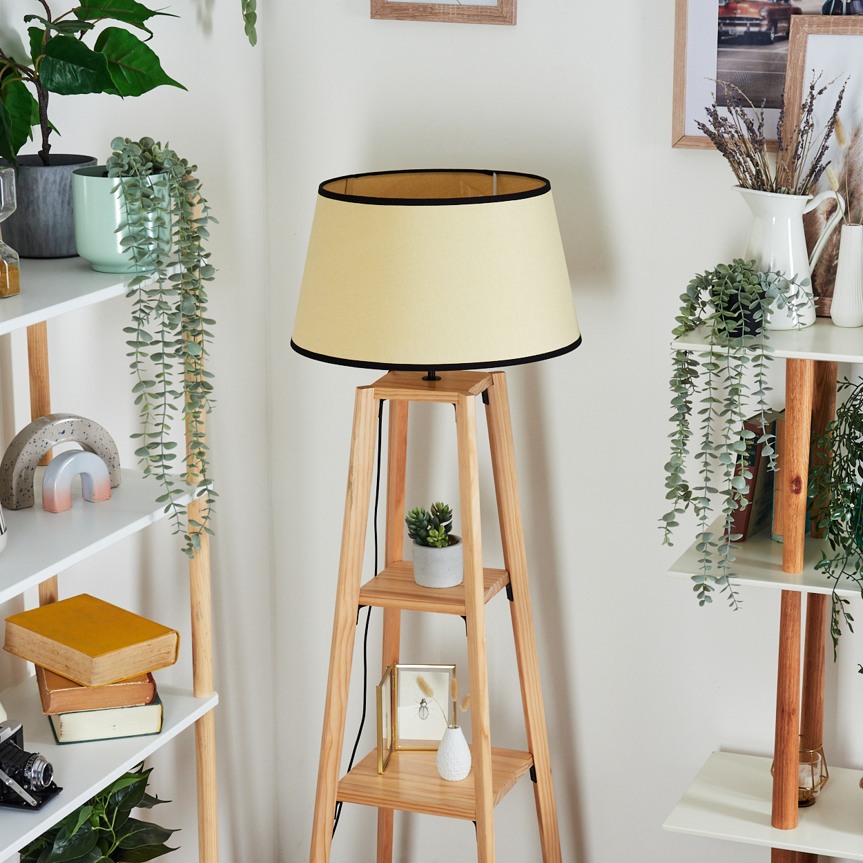 Eine Holzlampe mit beigem Schirm steht in der Nähe von Regalen mit Pflanzen und Büchern. Im Hintergrund sind gerahmte Bilder und weiße Wände zu sehen.