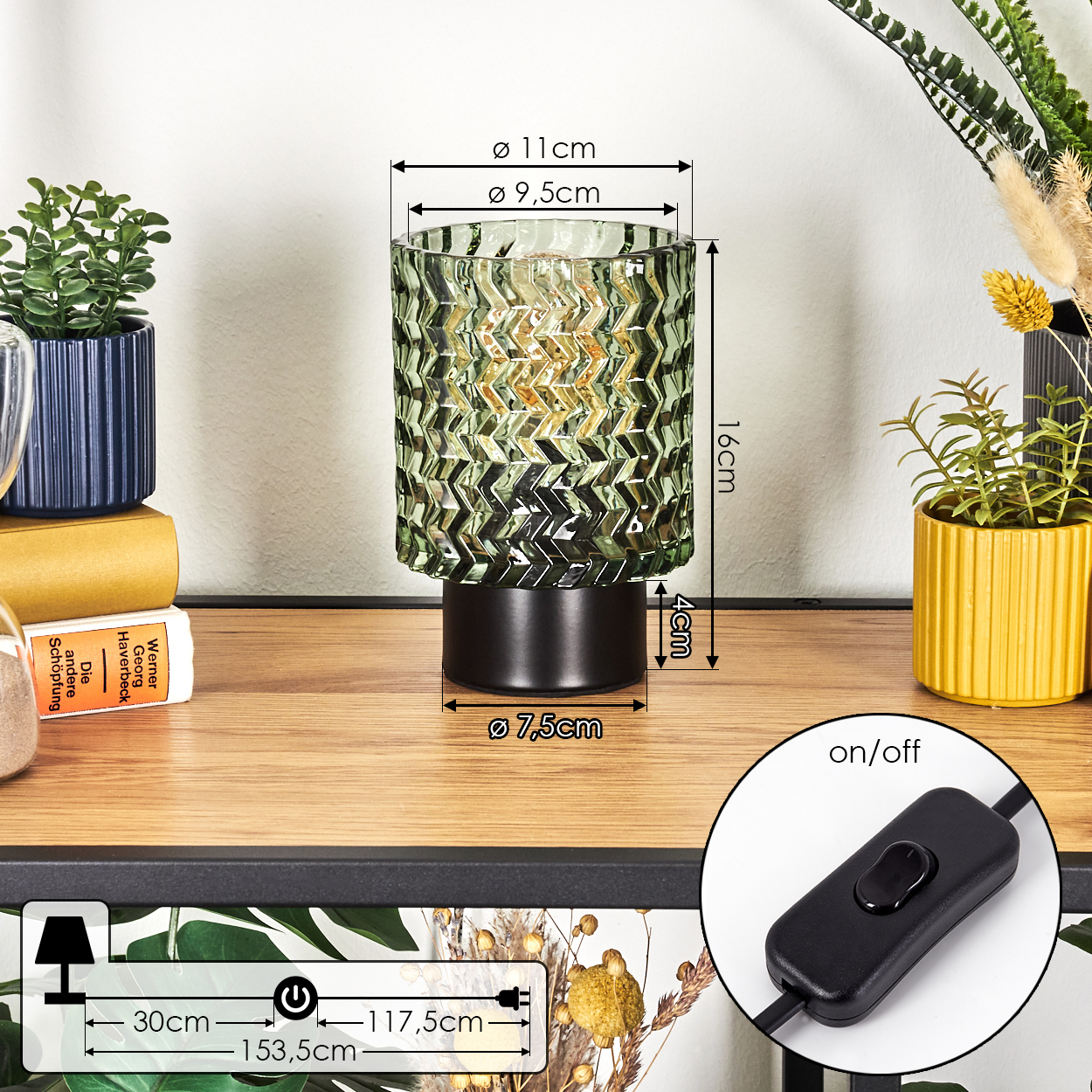 Eine dekorative Lampe mit grün gemustertem Glasschirm steht auf einer Holzoberfläche. Auch andere Pflanzen und ein Schalter sind sichtbar.