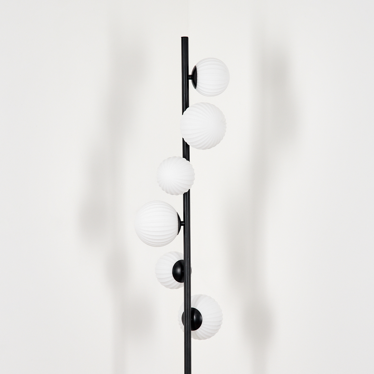 Eine schwarze Stehlampe mit sechs weißen Kugelleuchten. Die Kugeln haben ein strukturiertes Muster. Der Hintergrund ist eine schlichte weiße Wand.