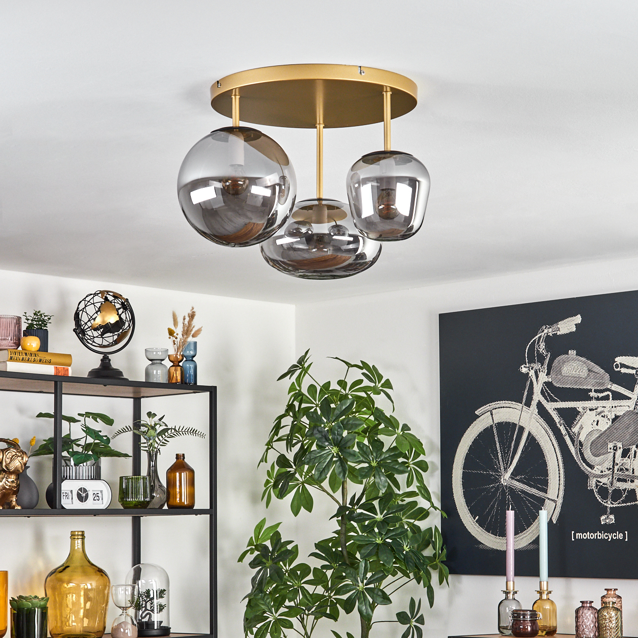 Eine moderne Deckenleuchte mit drei rauchigen Glaskugeln hängt über einem Raum mit einem Regal und einem Gemälde eines Motorrads.