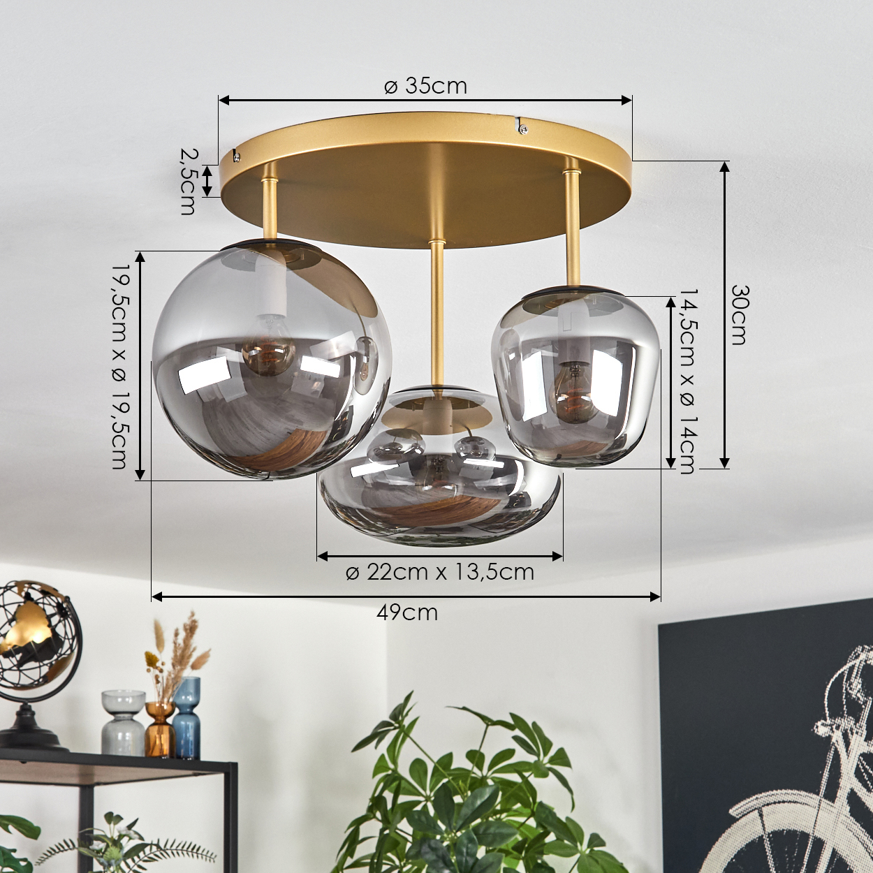 Eine moderne Deckenleuchte mit drei Rauchglaskugeln, goldenen Akzenten und beschrifteten Abmessungen. Sie hängt über einem Raum mit Dekoration und Pflanzen.