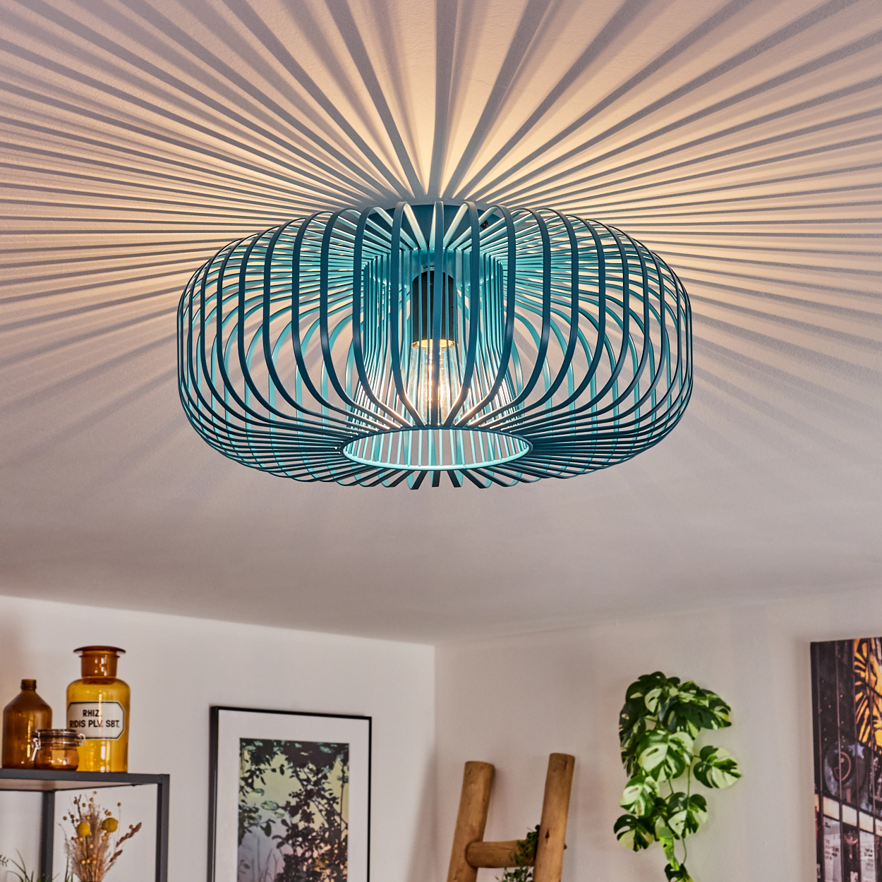 Eine türkisfarbene, käfigartige Deckenlampe wirft radiale Schatten. Darunter befinden sich Dekorationselemente wie eine Pflanze, Kunstwerke und Apothekergläser.