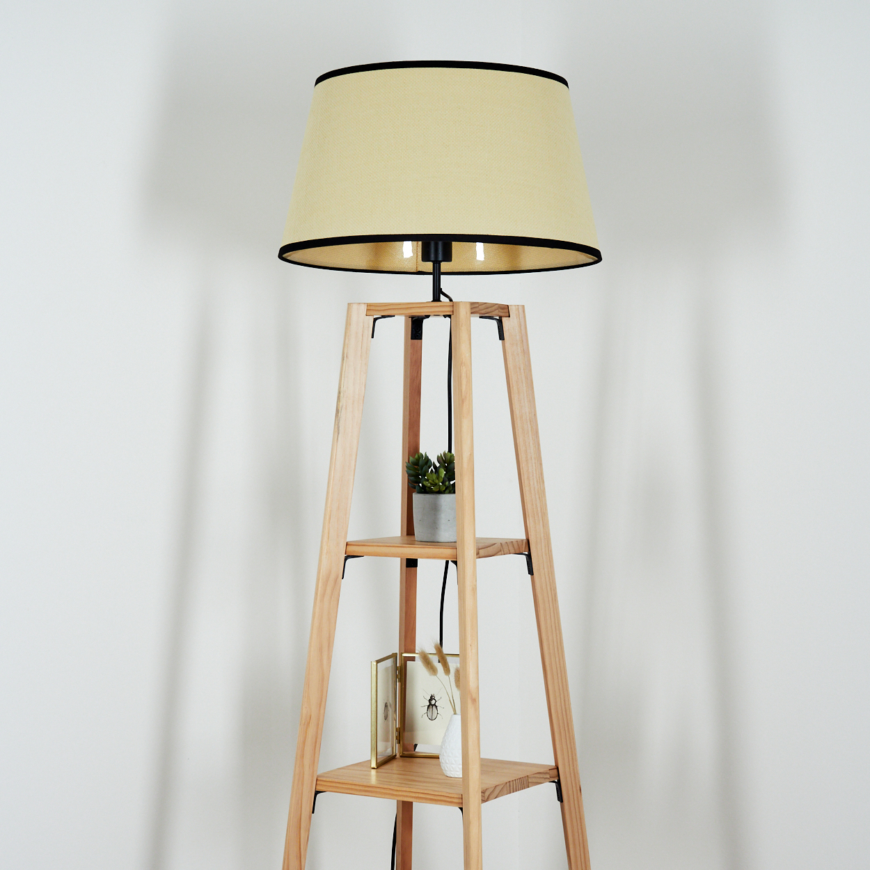Eine Stehlampe aus Holz mit beigem Lampenschirm, einer kleinen Pflanze und Dekorationsartikeln auf den Regalen. Der Hintergrund ist eine weiße Wand.