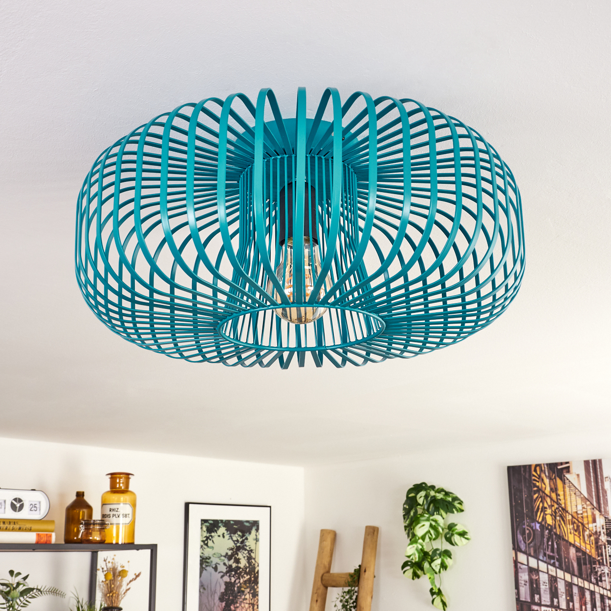 Eine türkisfarbene, käfigartige Deckenlampe mit sichtbarer Glühbirne. Hintergrund: weiße Decke, Dekoration und eine Leiter.