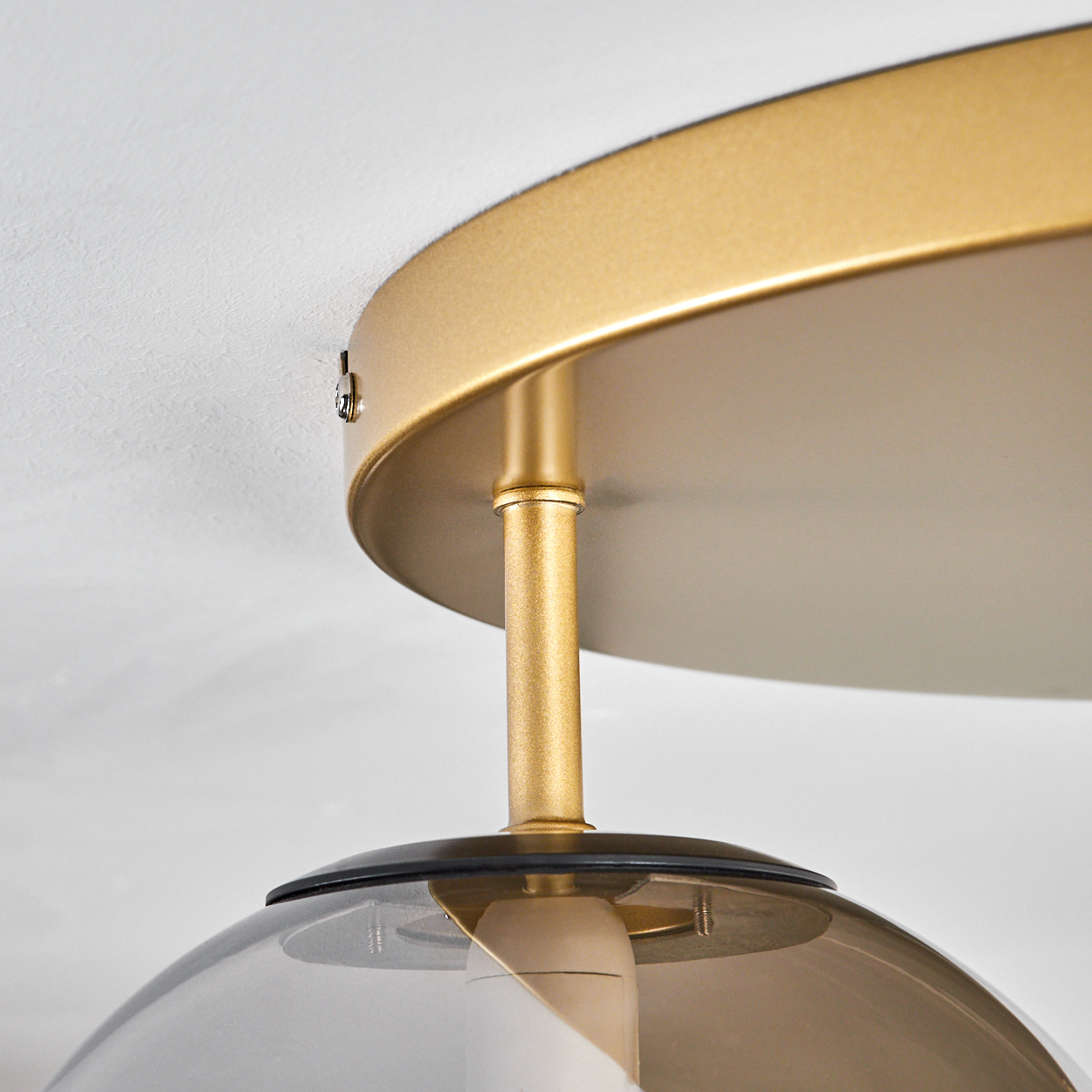 Nahaufnahme einer goldfarbenen Deckenleuchte. Die Leuchte hat einen runden Schirm und eine Glasabdeckung. Der Hintergrund ist eine weiße Decke.