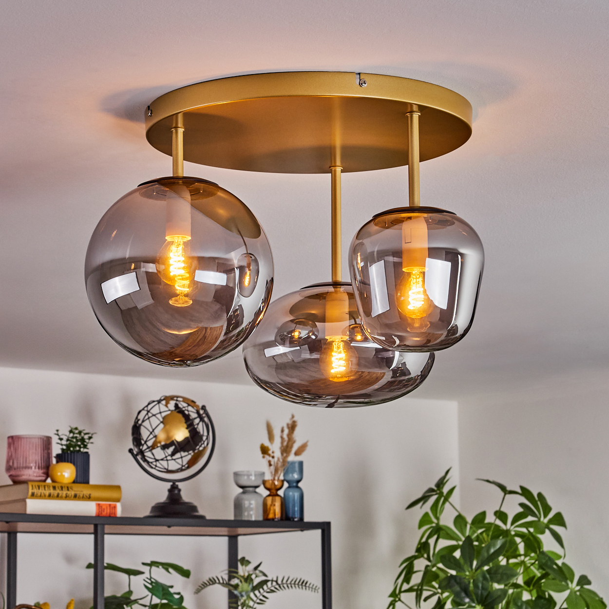 Eine Deckenleuchte mit drei rauchfarbenen Glaskugeln, goldenen Akzenten und sichtbaren Glühbirnen. Hintergrund: Regale und Pflanzen.