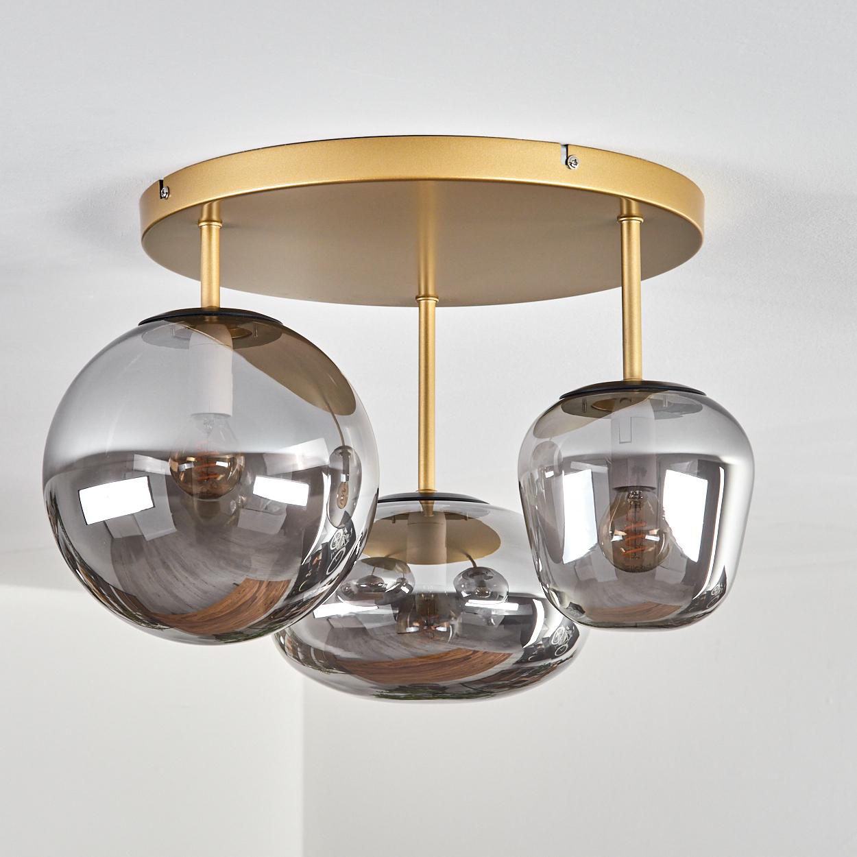 Eine Deckenleuchte mit drei grauen Glaskugeln, die von einer goldenen kreisförmigen Basis hängen. Die Glühbirnen sind in den Kugeln sichtbar.