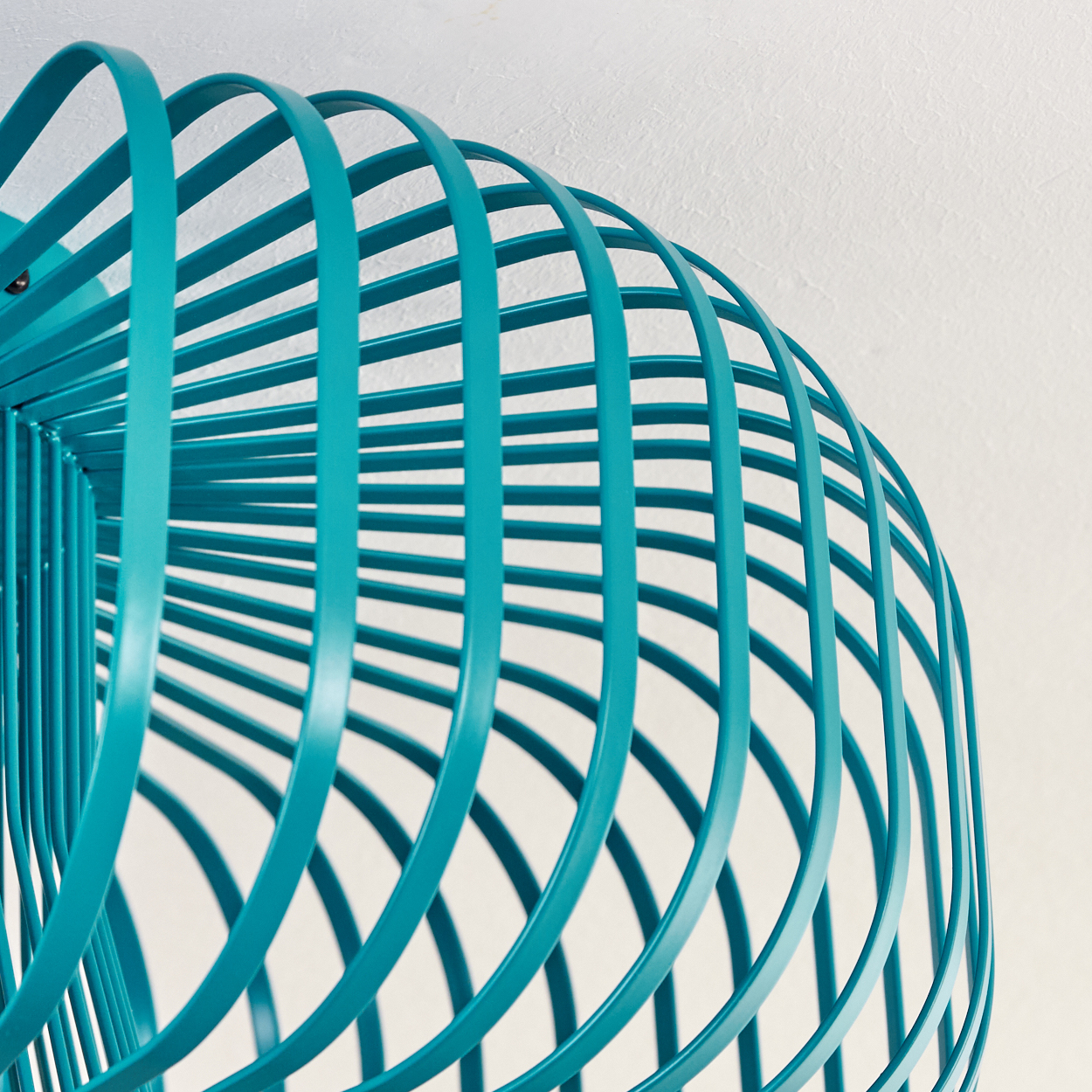 Nahaufnahme einer teal-farbenen, käfigartigen Struktur vor einer weißen Wand. Die Struktur besteht aus gebogenen Metallstäben.