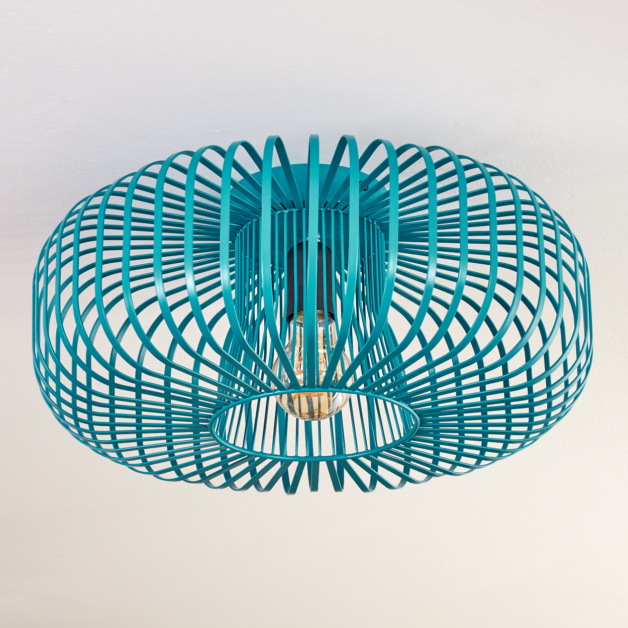 Eine türkisfarbene, käfigartige Deckenlampe. Die Glühbirne ist im Inneren sichtbar. Der Hintergrund ist eine schlichte weiße Decke. Die Lampe ist zentriert.