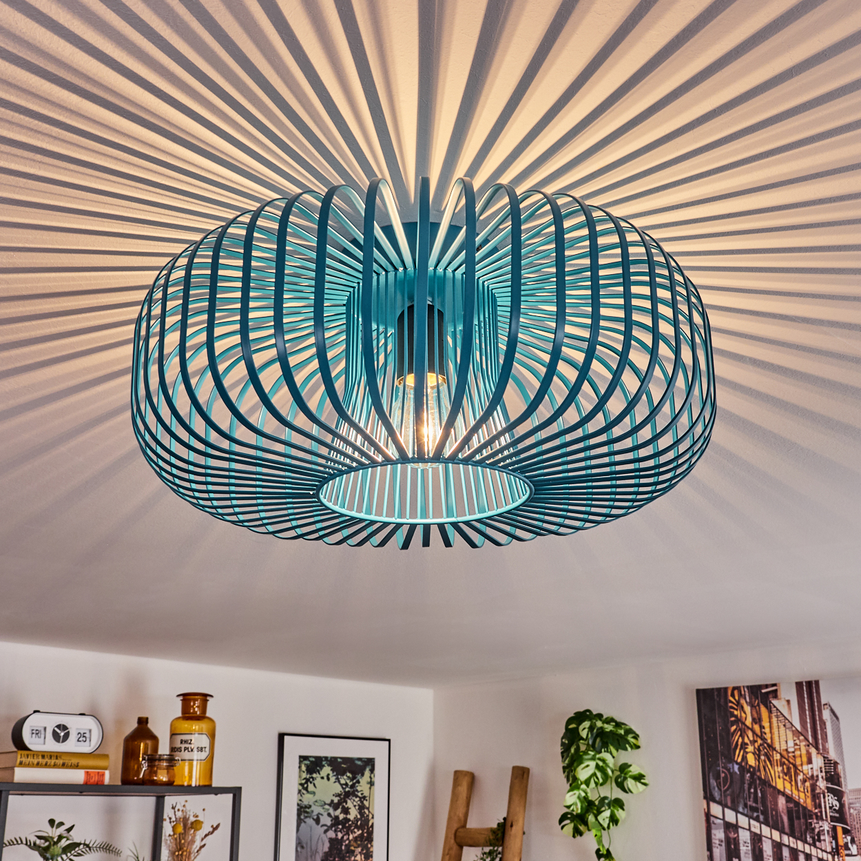 Eine teal-farbene, käfigartige Deckenlampe ist das Hauptaugenmerk. Sie wirft radiale Schatten auf die weiße Decke. Bücher, Kunst und Pflanzen stehen auf einem Regal darunter.