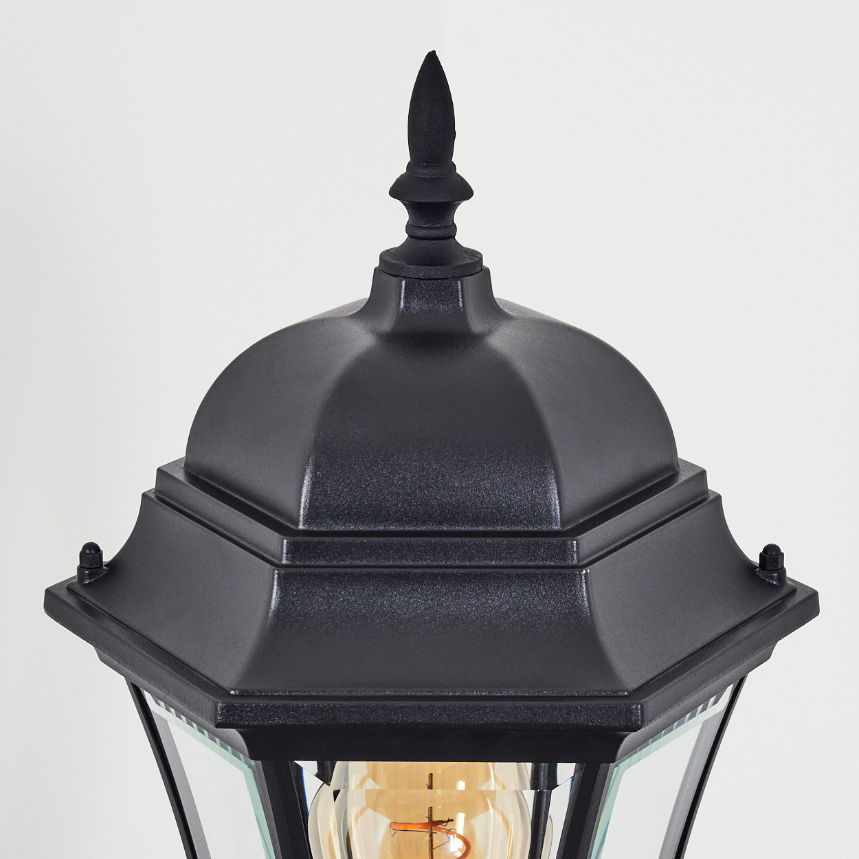 Nahaufnahme einer schwarzen Außenlampe. Sie hat eine dekorative Oberseite und eine gläserne Glühbirne im Inneren. Der Hintergrund ist weiß.