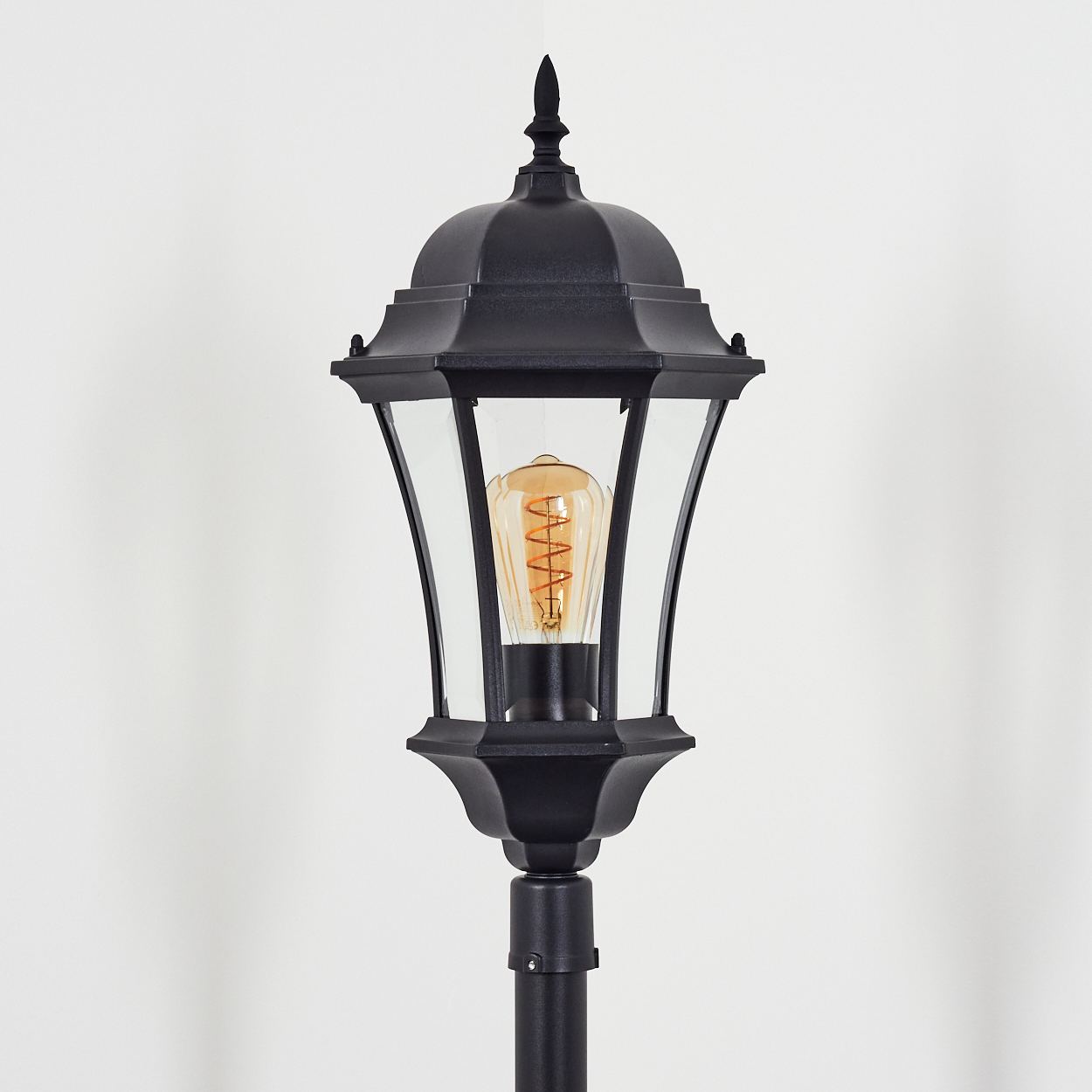 Schwarze Außenlampe mit Glaspaneelen und dekorativer Spitze, mit einer warm getönten Glühbirne vor weißem Hintergrund.