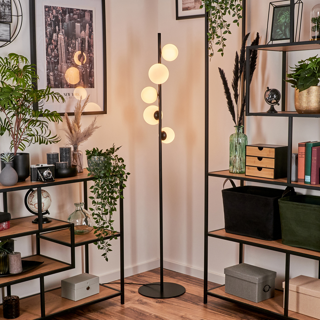 Ein Wohnzimmer mit einer Stehlampe mit Kugelleuchten. Regale mit Dekor und Pflanzen befinden sich auf beiden Seiten. Ein NYC-Poster hängt an der Wand.