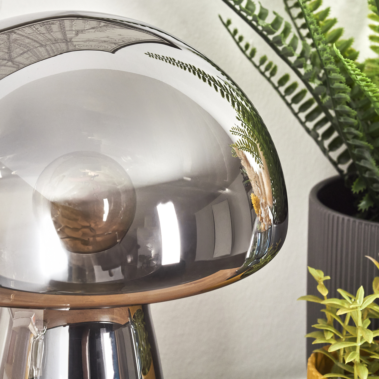 Eine pilzförmige Lampe mit einem reflektierenden grauen Schirm. Im Hintergrund sind grüne Pflanzen in Töpfen zu sehen, die sich in der Lampe spiegeln.