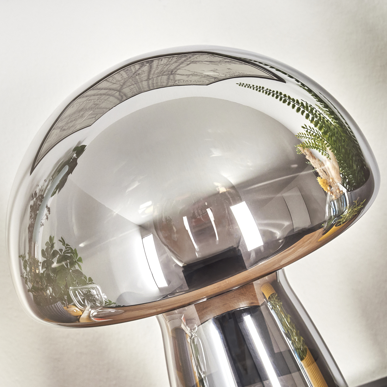Eine pilzförmige Lampe mit reflektierendem Glasschirm. Der Schirm reflektiert Pflanzen und einen Raum. Der Sockel ist aus Holz und Metall.