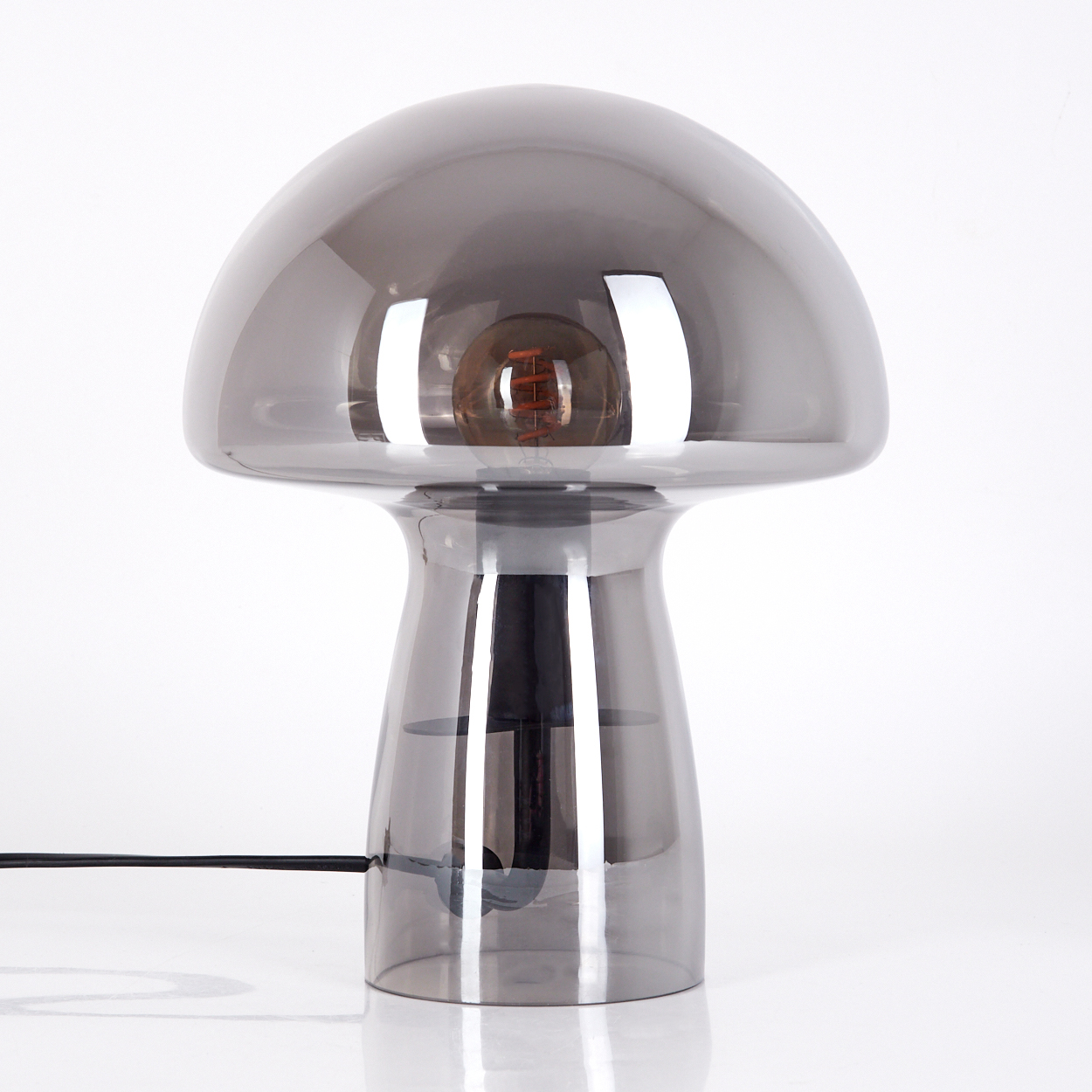 Graue pilzförmige Lampe. Sie hat eine Glühbirne. Sie steht auf einer weißen Oberfläche. Ein schwarzes Kabel geht von der Basis aus.