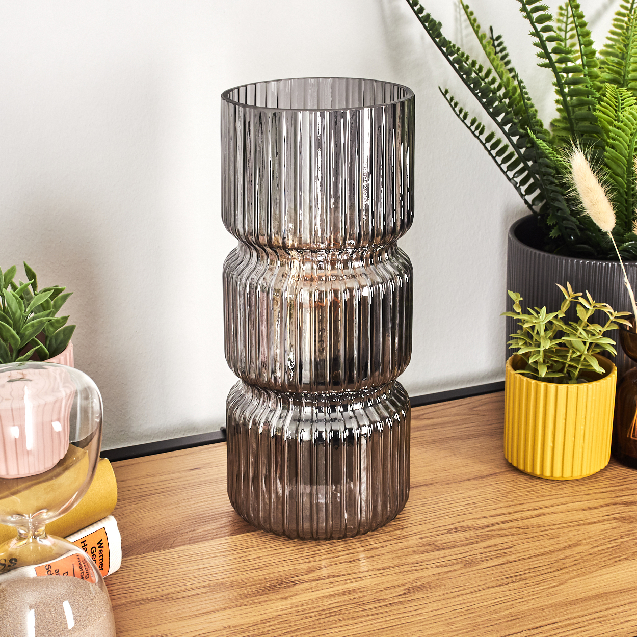 Eine gerippte Glasvase steht auf einer Holzoberfläche. Pflanzen und eine Sanduhr sind im Hintergrund. Die Vase reflektiert Licht.