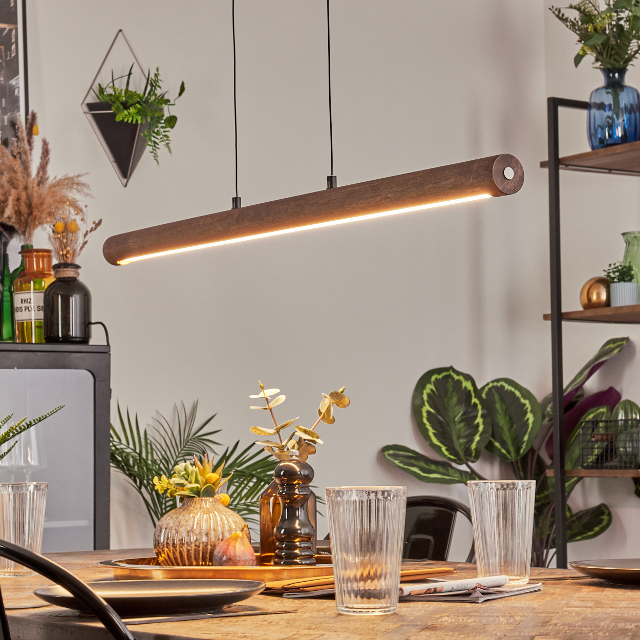 Ein Esszimmer mit einer Holzleuchte, einem Tisch mit Gläsern und Dekor sowie Pflanzen im Hintergrund.