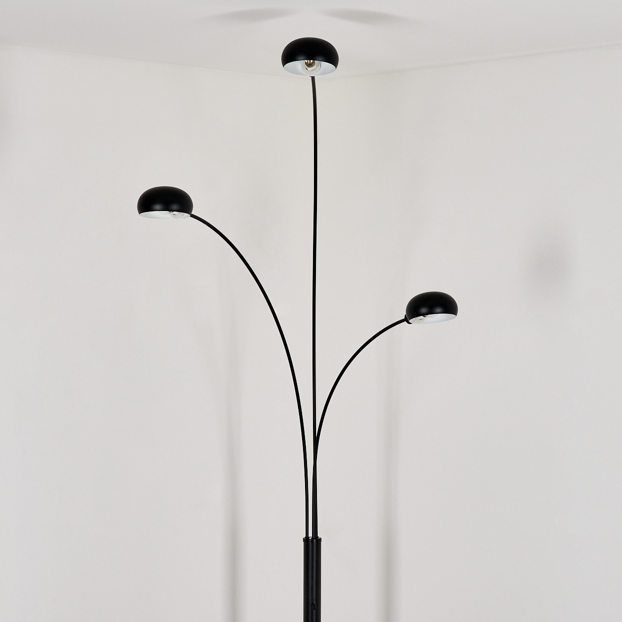Eine schwarze Stehlampe mit drei Köpfen steht vor einer weißen Wand. Die Lampe hat drei gebogene Arme mit schwarzen Lampenschirmen.