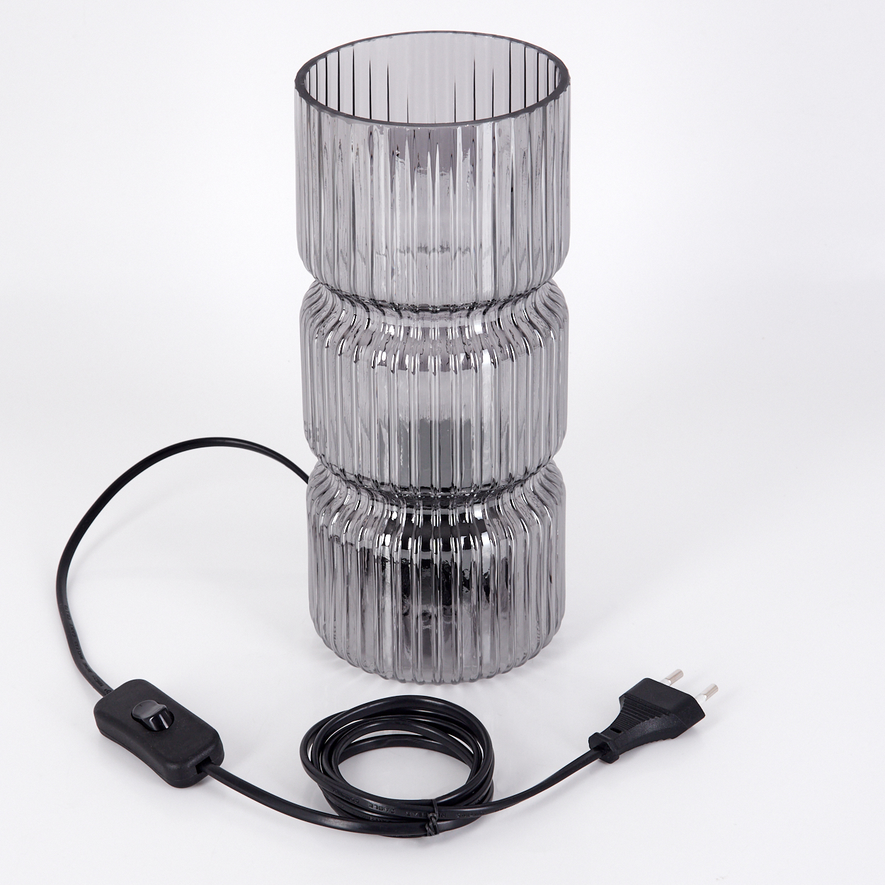 Eine graue Glaslampe mit schwarzem Netzkabel und Stecker steht auf einer weißen Oberfläche. Die Lampe hat ein geripptes Design.