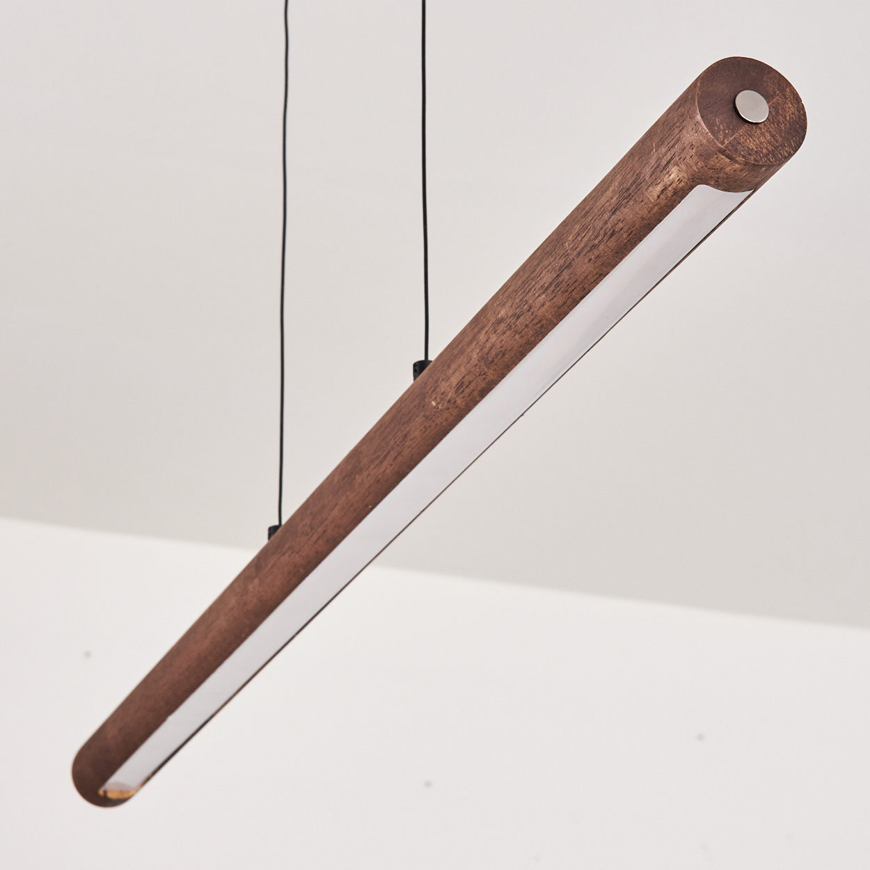 Eine zylindrische Holzlampe hängt an schwarzen Drähten vor weißem Hintergrund. Die Lampe hat einen weißen Lichtstreifen.