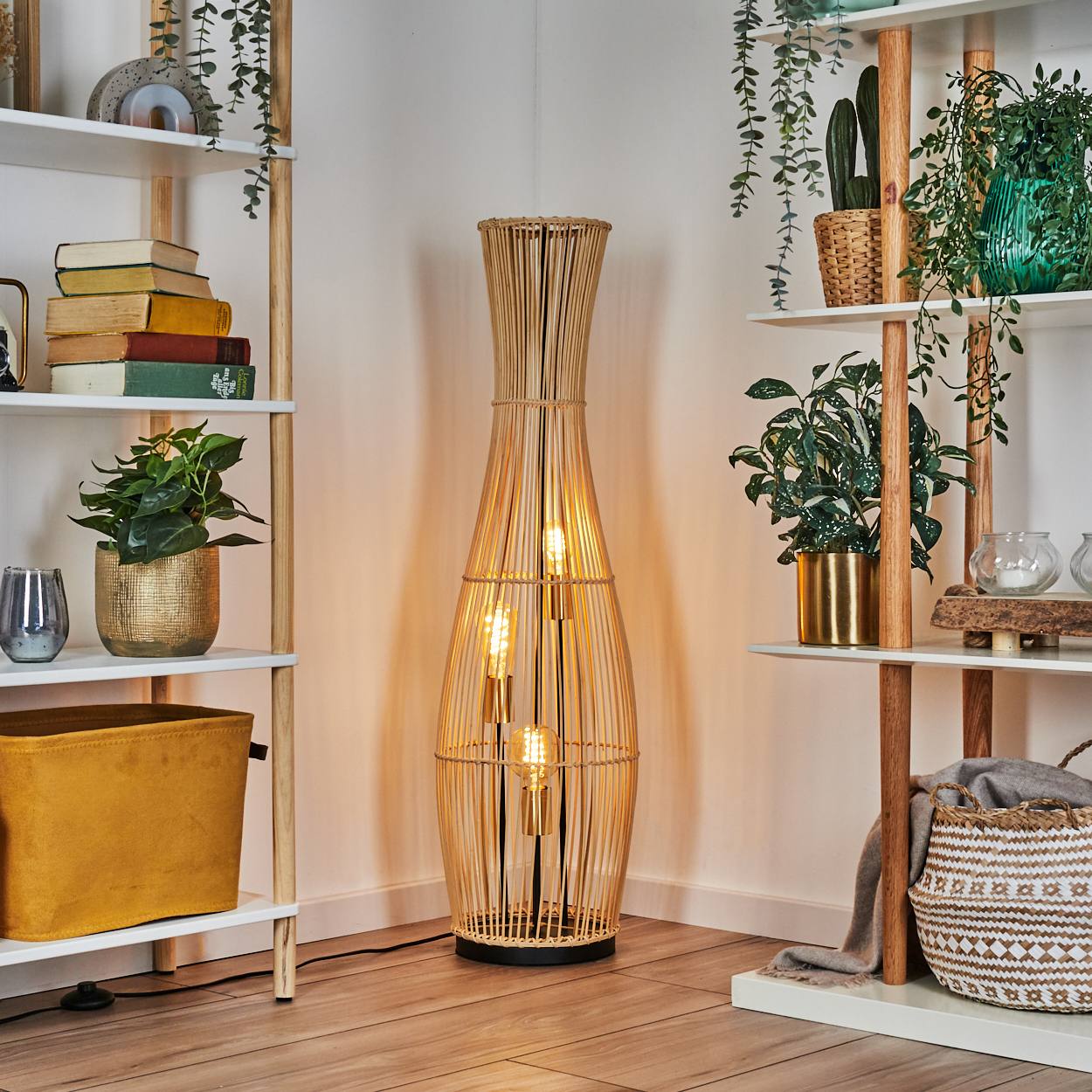 Eine hohe, gewebte Lampe steht zwischen zwei weißen Regalen mit Pflanzen und Dekoration. Holzböden und eine weiße Wand vervollständigen die Szene.
