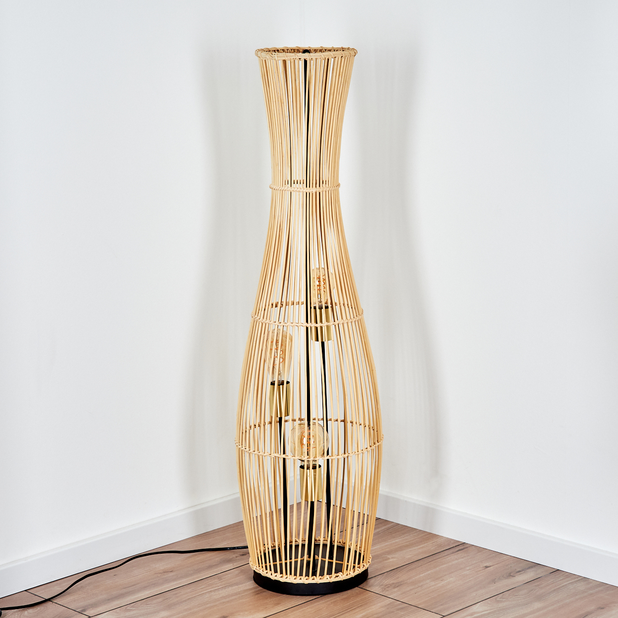 Eine hohe, gewebte Stehlampe aus Bambus steht in einer Ecke. Sie hat einen schwarzen Sockel, drei Glühbirnen und steht vor einer weißen Wand und Holzboden.