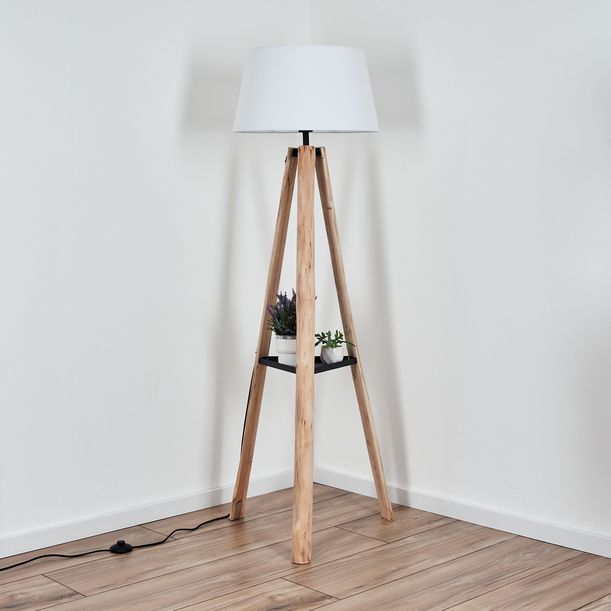 Eine hölzerne Stativ-Stehlampe mit weißem Lampenschirm steht in einer Ecke. Sie hat ein Regal mit Pflanzen. Der Boden ist aus Holz.
