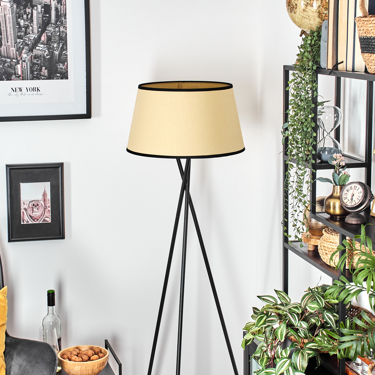 Eine Stehlampe mit beigefarbenem Schirm und schwarzem Gestell steht neben einem Bücherregal mit Dekoration und Pflanzen. Ein New York-Druck hängt an der Wand.