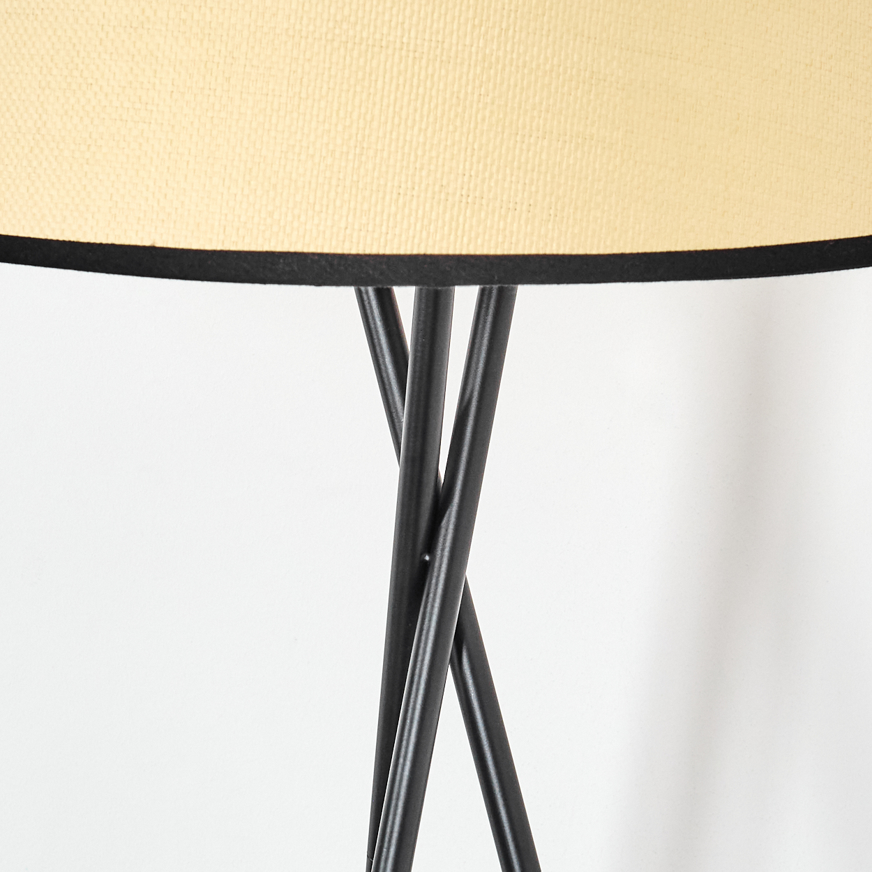 Nahaufnahme einer Lampe. Der Lampenschirm ist cremefarben mit schwarzem Rand. Schwarze Metallbeine tragen die Lampe vor einer weißen Wand.