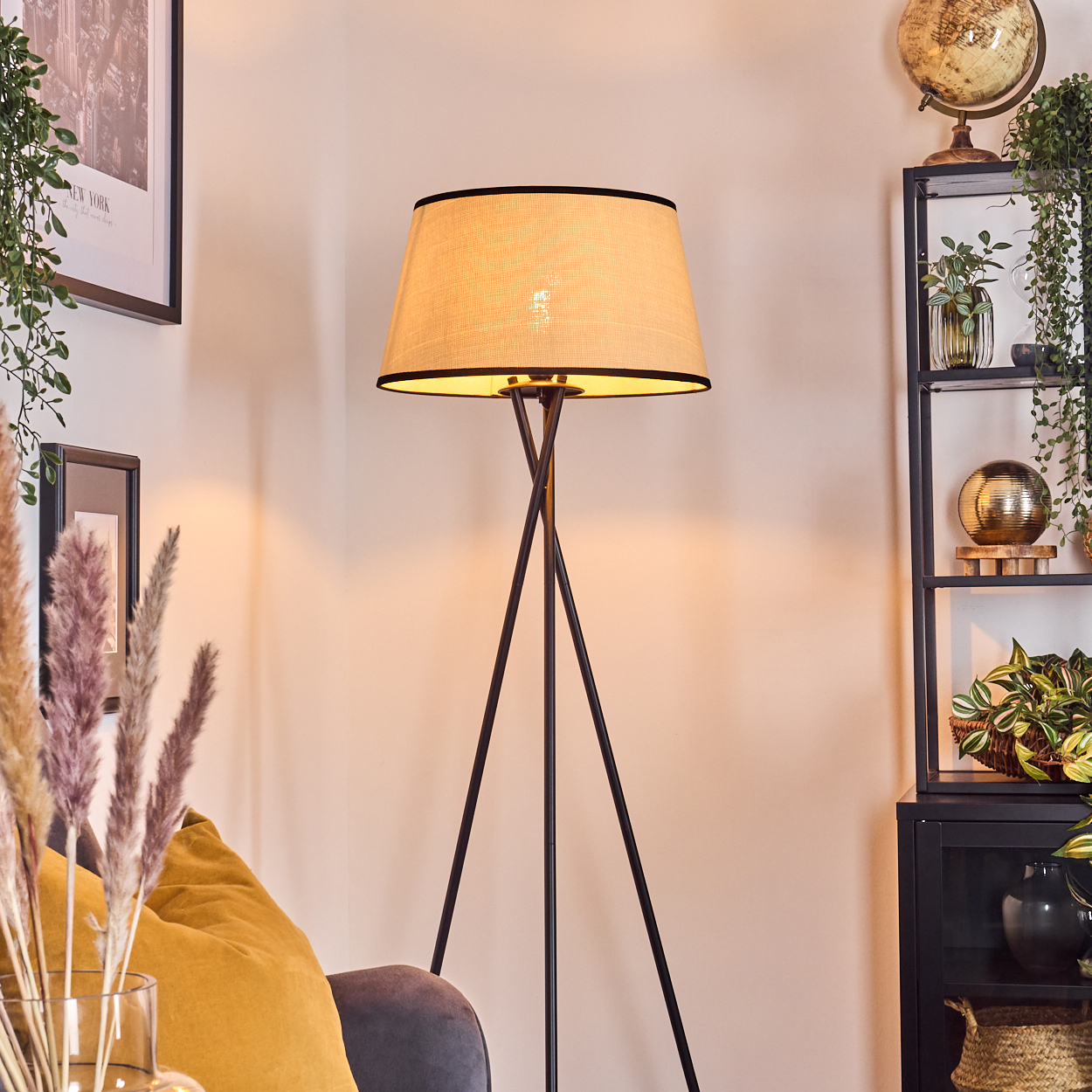 Eine beleuchtete Stehlampe mit beigem Schirm steht in einem Raum. Im Hintergrund sind ein Regal mit Pflanzen und ein Globus zu sehen.
