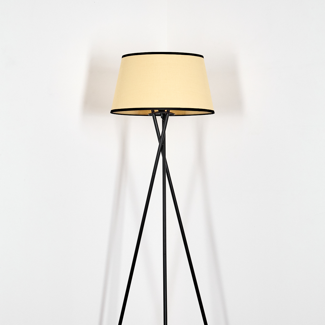 Eine Stehlampe steht vor einer weißen Wand. Die Lampe hat einen beigefarbenen Schirm mit schwarzem Rand und ein schwarzes Stativ.
