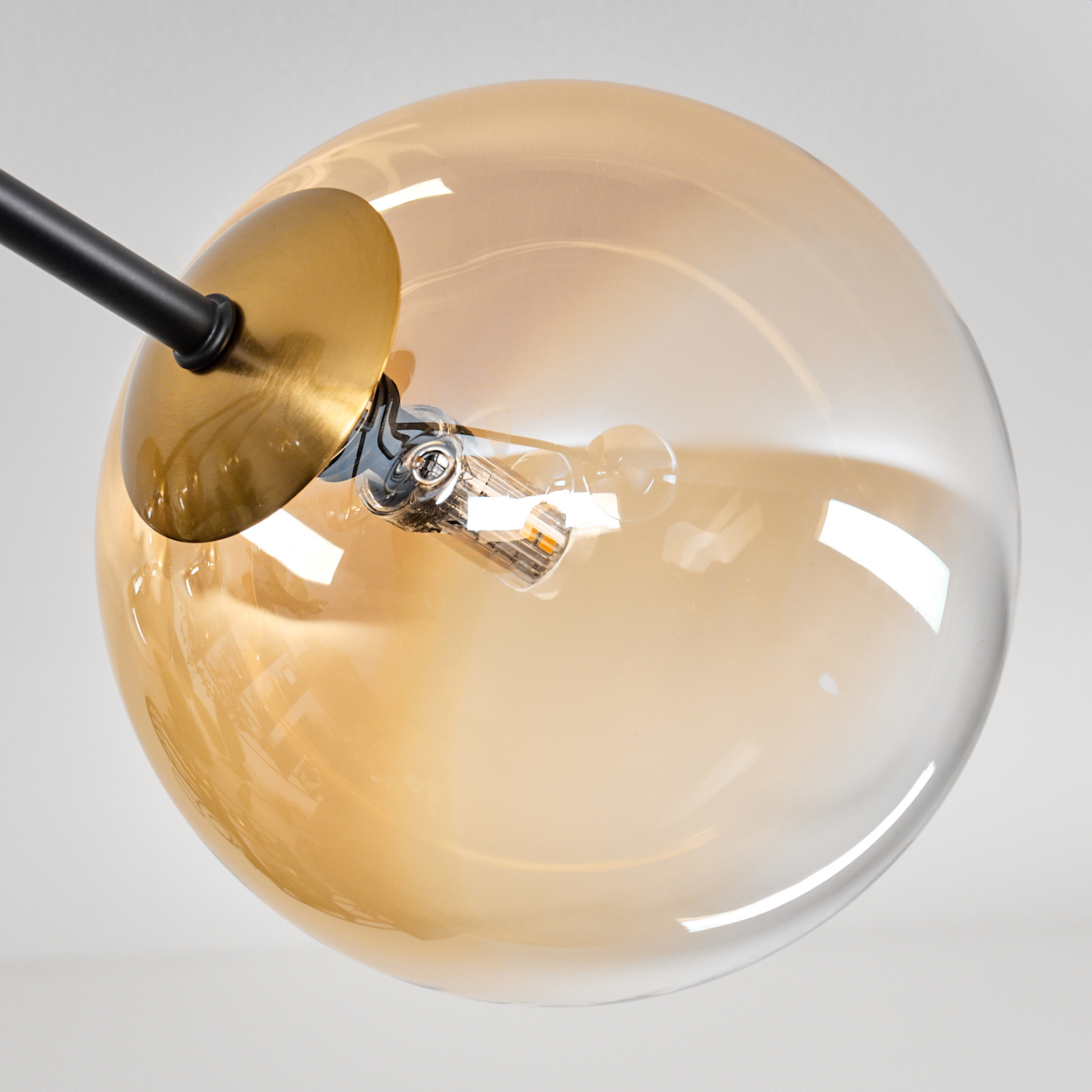 Nahaufnahme einer Leuchte. Ein klarer Glaskolben mit Goldtönung umgibt eine Glühbirne. Schwarzer Metallarm und eine goldene Platte.