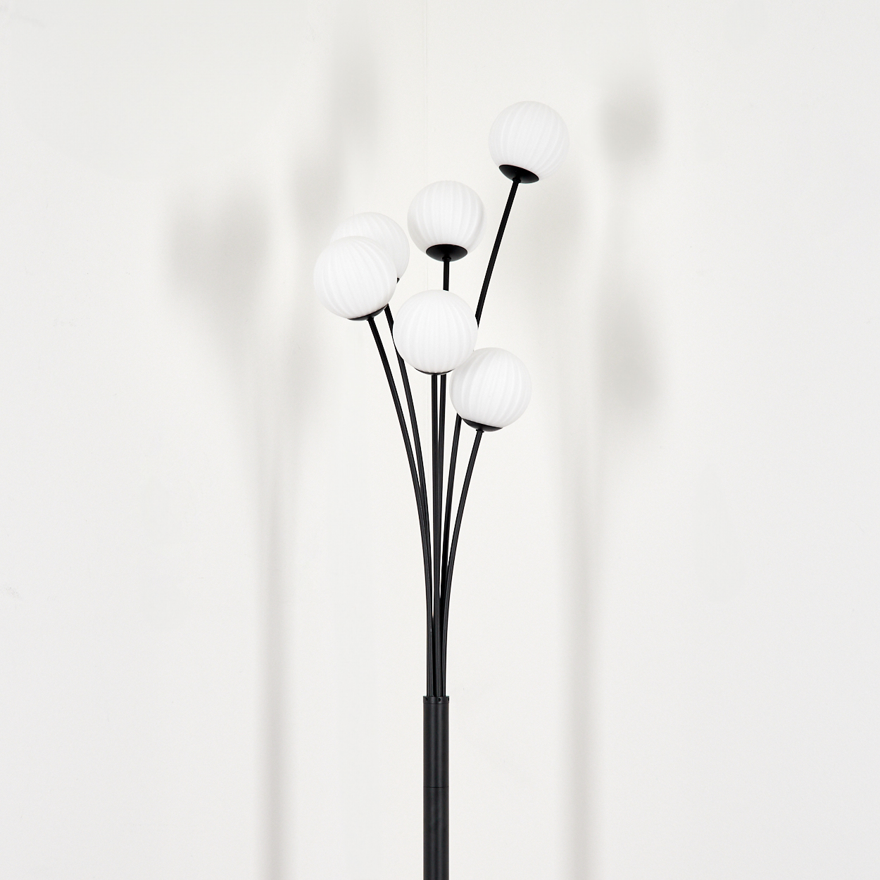 Eine schwarze Stehlampe mit sechs weißen Kugelleuchten. Der Hintergrund ist eine weiße Wand mit dem Schatten der Lampe.