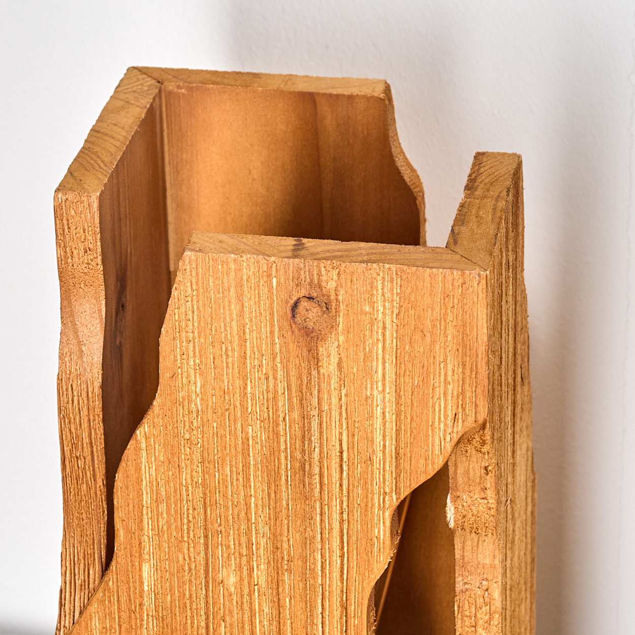 Nahaufnahme einer Holzkonstruktion. Das Holz ist hellbraun mit sichtbarer Maserung. Die Struktur hat eine unregelmäßige Form und befindet sich vor einer weißen Wand.