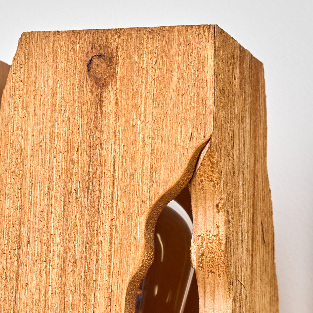 Nahaufnahme einer Holzstruktur mit einem geschwungenen Ausschnitt, der eine Flasche im Inneren freilegt. Das Holz ist hellbraun, vor weißem Hintergrund.