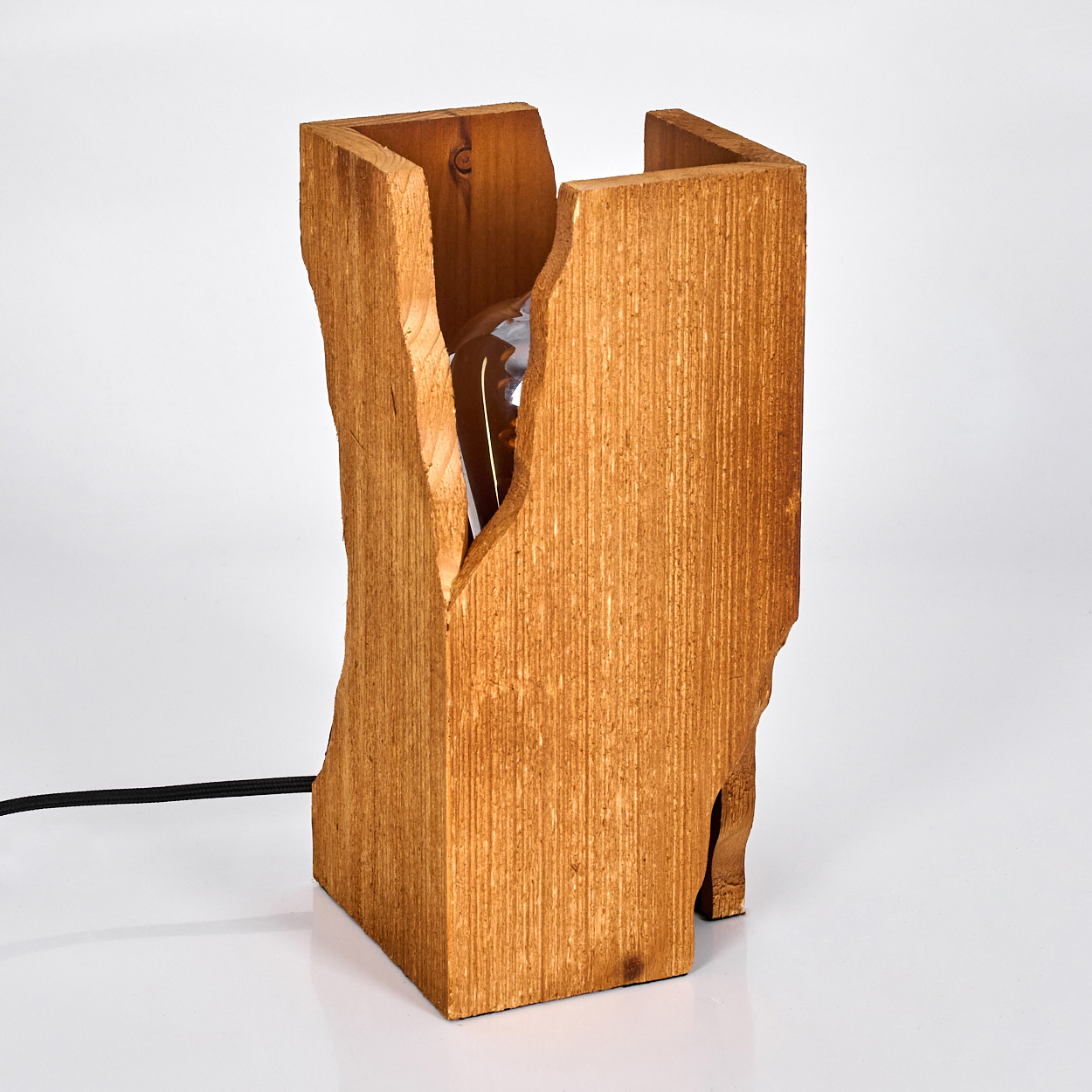 Eine Holzlampe mit einer Glühbirne. Die Lampe hat eine rechteckige Form mit rauen Kanten. Ein schwarzes Kabel ist befestigt.