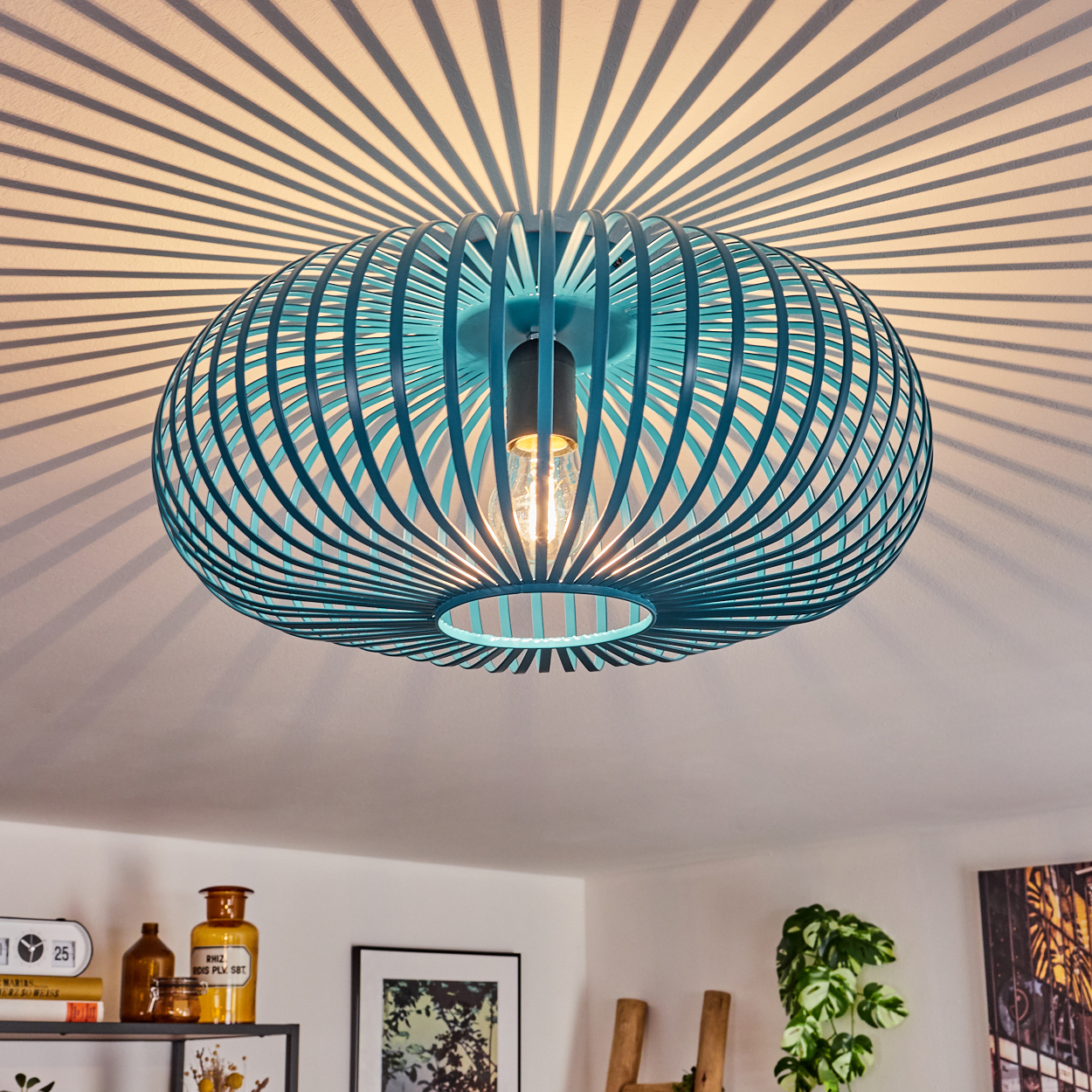 Eine türkisblaue, käfigartige Deckenlampe mit Glühbirne, die Schattenmuster an die Decke wirft. Regale und eine Pflanze sind darunter sichtbar.
