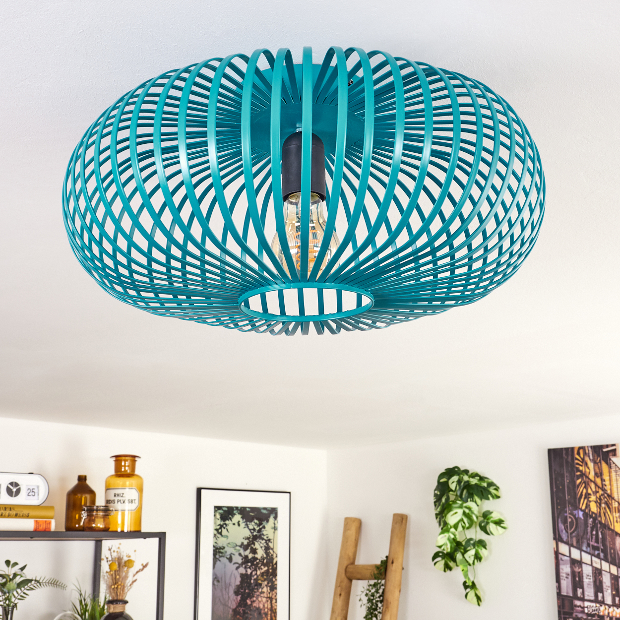 Eine teal-farbene, kugelförmige Deckenlampe mit sichtbarer Glühbirne. Im Hintergrund sind Regal, Kunst, Leiter und Pflanzen zu sehen.